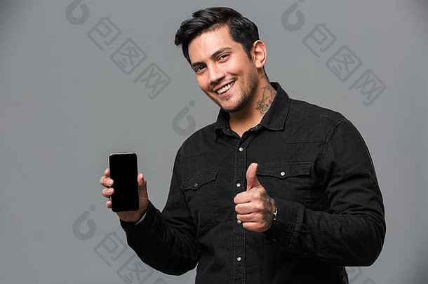 照片微笑年轻的英俊的男人。站孤立的灰色墙相机显示拇指显示移动电话