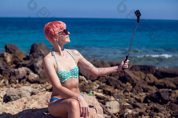 旅行休闲夏天技术人概念年轻的女人粉红色的短头发采取自拍海滩背景