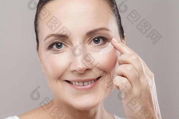 中间岁的女人完美的脸皮肤应用化妆品奶油皮肤眼睛相机露出牙齿的微笑孤立的灰色背景
