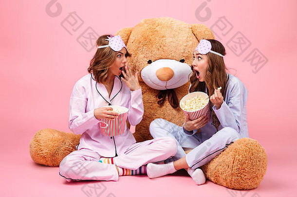震惊了漂亮的女孩穿着睡衣坐着大泰迪熊吃爆米花孤立的粉红色的背景