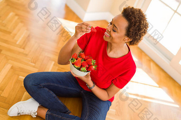 美丽的年轻的非洲女人非洲式发型头发吃新鲜的草莓坐着地板上