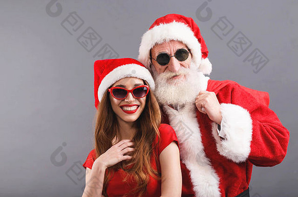 圣诞老人老人白色胡子穿sungasses年轻的夫人老人穿圣诞老人他红色的衣服太阳镜站笑灰色的背景一年圣诞节假期记忆礼物购物折扣商店雪少女圣诞老人老人化妆发型狂欢节