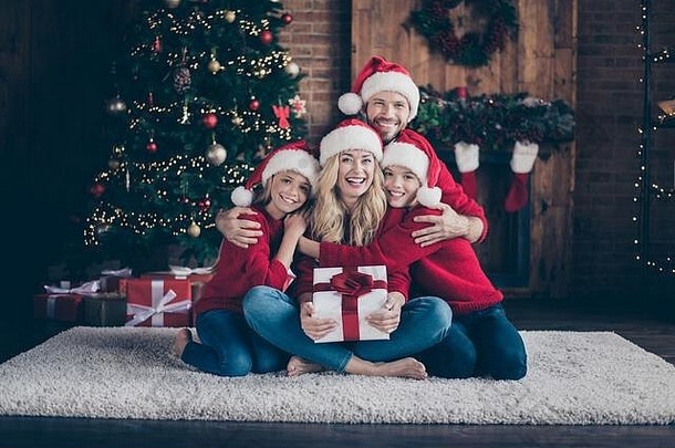 完整的成员家庭爸爸妈妈妹妹哥哥给惊喜礼品盒支出x-mas夏娃坐着地板上newyear树在室内穿圣诞老人帽