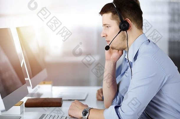 年轻的但耳机坐着桌子上阳光明媚的现代办公室听客户调用中心运营商工作