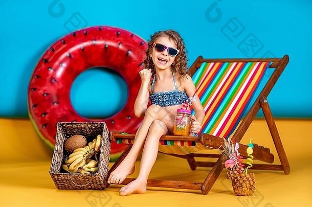 微笑女孩穿泳衣太阳镜持有Jar汁鸡尾酒五彩缤纷的吸管坐着彩虹甲板椅子大西瓜充气圆夏天童年概念