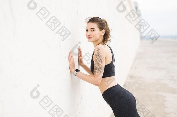 年轻的美丽的微笑女人黑暗灰色的运动前紧身裤幸福的相机倾斜墙练习户外
