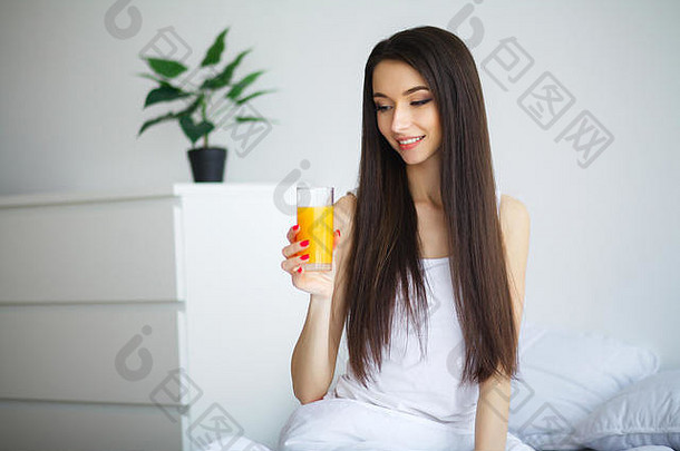 年轻的女人享受玻璃橙色汁早....放松坐着床上健康的饮食概念
