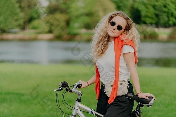 漂亮的女人游乐设施自行车浓密的金发女郎头发站自行车穿太阳镜白色衬衫享受旅行新鲜的空气小成河