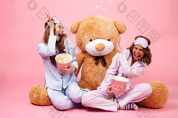 快乐漂亮的女孩穿着睡衣有趣的坐着坐着大泰迪熊吃爆米花孤立的粉红色的背景