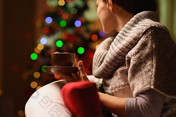 年轻的女人坐着椅子热饮料前面圣诞节树后视图