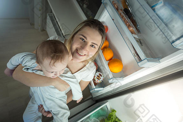 肖像年轻的妈妈。持有婴儿男孩站开放冰箱厨房晚上