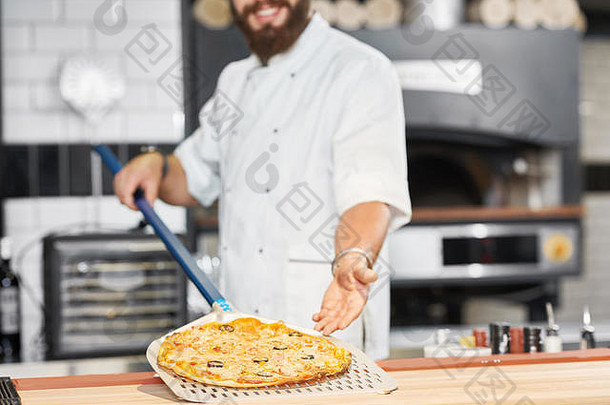 裁剪照片贝克持有新鲜的烤披萨铺设长金属铲微笑男人。穿白色酋长束腰外衣工作空间餐厅厨房大烤箱设备