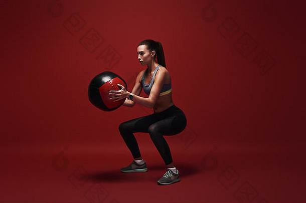 美丽的女运动员下蹲锻炼球站红色的背景