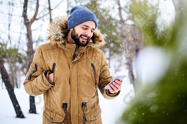 微笑有胡子的男人。穿温暖的冬天衣服智能手机快互联网数据连接国家一边英俊的男人。发短信手机应用程序森林降雪森林