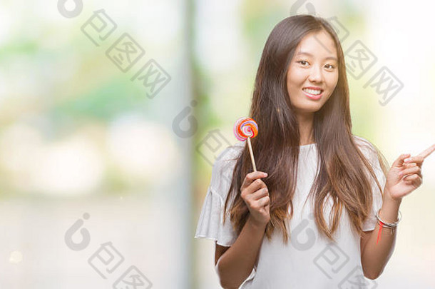 年轻的亚洲女人吃棒棒糖糖果孤立的背景快乐指出手手指一边