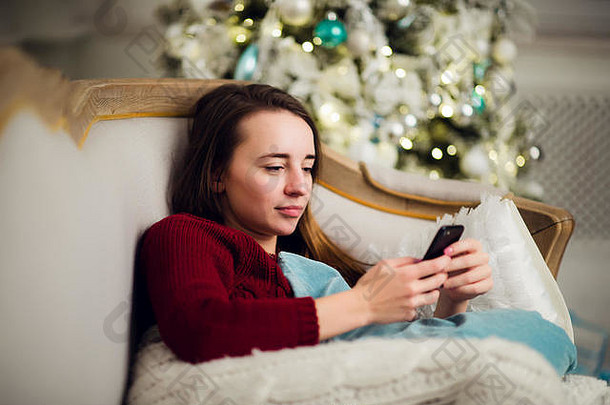 年轻的女人检查文本消息移动电话liyng沙发前面圣诞节树首页