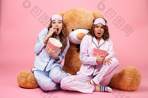 害怕漂亮的女孩穿着睡衣坐着大泰迪熊吃爆米花孤立的粉红色的背景
