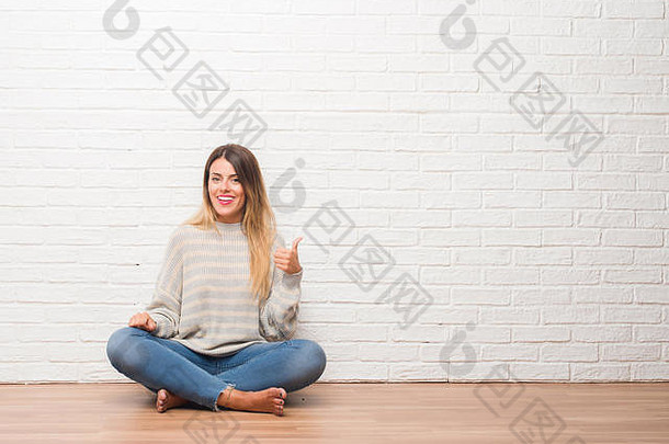 年轻的成人女人坐着<strong>地板</strong>上白色砖墙首页微笑快乐脸指出一边拇指