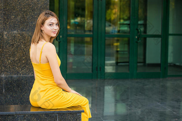 美丽的女人坐着把回来相机摆姿势女孩站在户外绿色建筑使玻璃黄色的跳