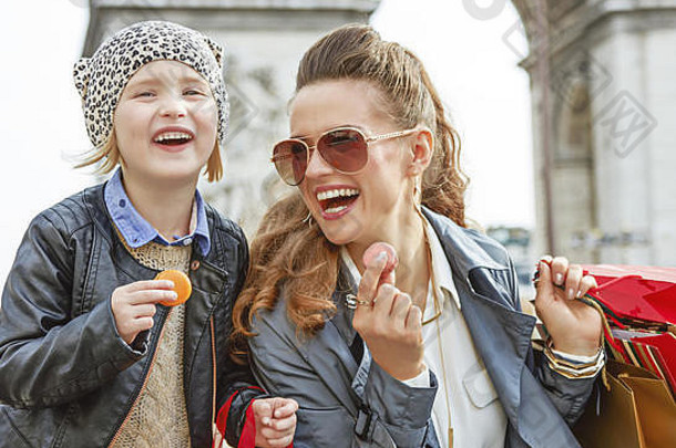 时尚的秋天巴黎微笑时尚的妈妈。女儿购物袋弧凯旋门巴黎法国吃蛋白杏仁饼
