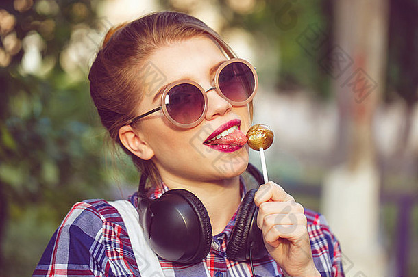 年轻的赶时髦的人女孩听音乐耳机夏天公园特写镜头肖像女孩舔棒棒糖温暖的爽肤水