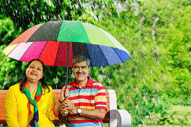 快乐上了年纪的夫妇坐着板凳上伞雨保护在花园