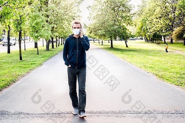 年轻的成人高加索人男人。穿保护脸面具走持有电话会说话的户外生活风格冠状病毒流感大流行概念