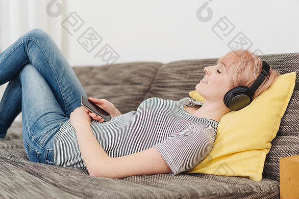 快乐年轻的女人说谎舒适的沙发支持缓冲听音乐立体声耳机幸福的微笑