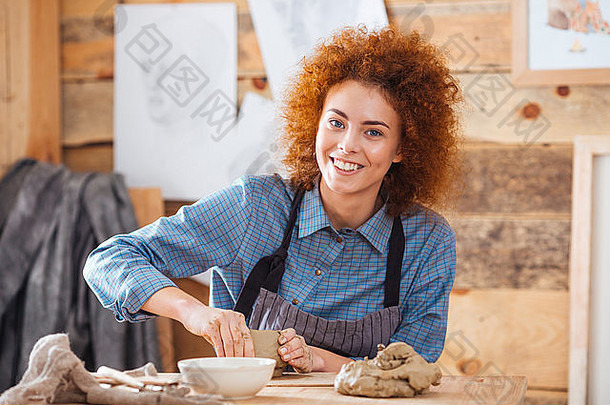 快乐的年轻的女人波特卷曲的红色的头发围裙坐着工作艺术陶器工作室