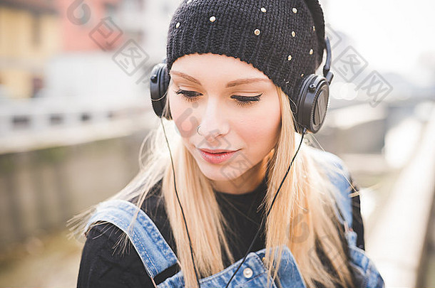 一半长度年轻的美丽的金发女郎直头发女人城市耳机听音乐向下沉思的深思熟虑的放松技术概念