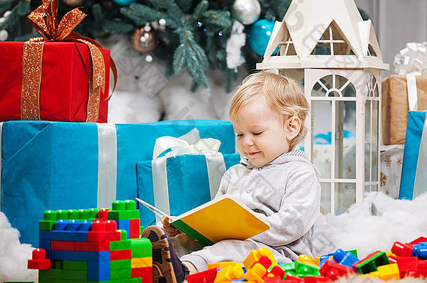 可爱的蹒跚学步的男孩坐着圣诞节树阅读书建筑块分散