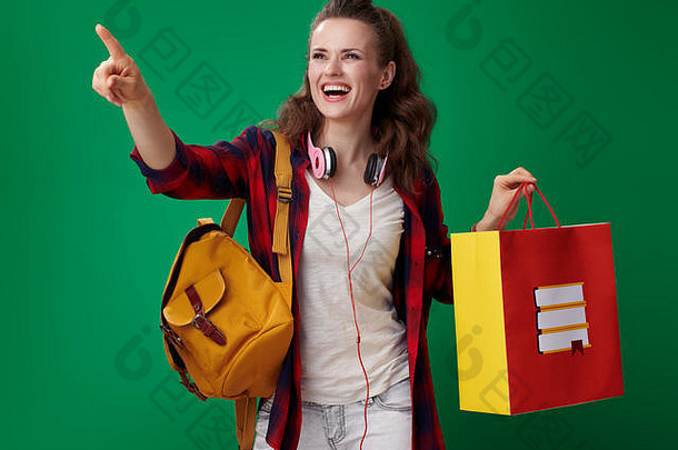 微笑年轻的学生女人红色的衬衫背包耳机持有购物袋书指出绿色背景