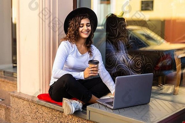 美丽的女人坐着板凳上持有咖啡移动PC户外
