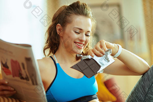 快乐活跃的体育女人健身衣服现代房子阅读杂志吃巧克力