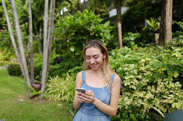 年轻的高加索人女人聊天智能手机花园树穿牛仔裤背心裙