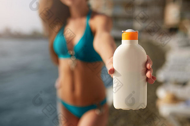 快乐女人显示晒黑防晒霜奶油白色瓶模糊海滩背景晒黑苗条的女孩穿蓝色的比基尼泳衣持有