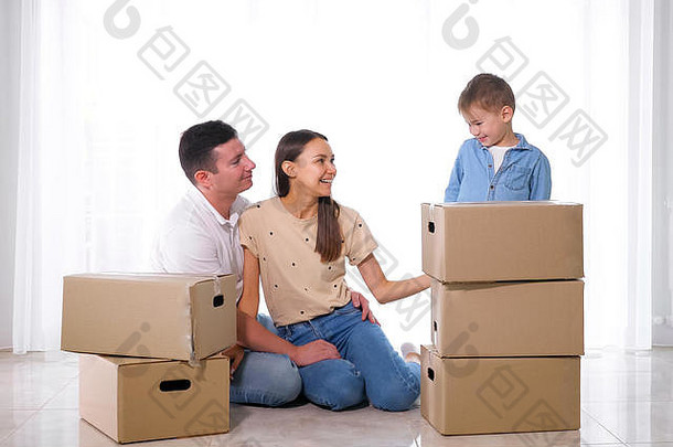快乐小儿子站大纸板盒子父母坐着光地板上房子