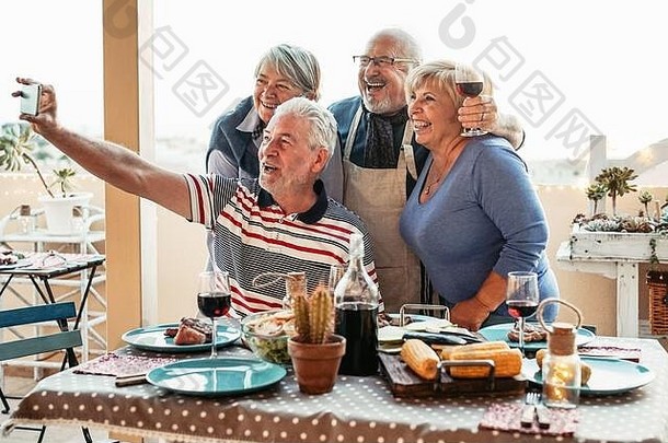 快乐老年人朋友采取自拍移动智能手机相机晚餐阳台退休人有趣的吃喝红色的酒