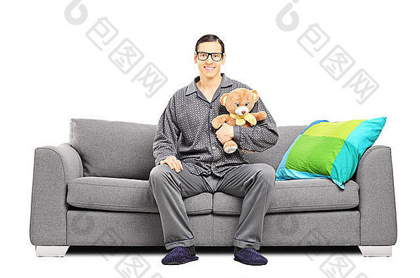 年轻的男人。睡衣坐着沙发持有泰迪熊