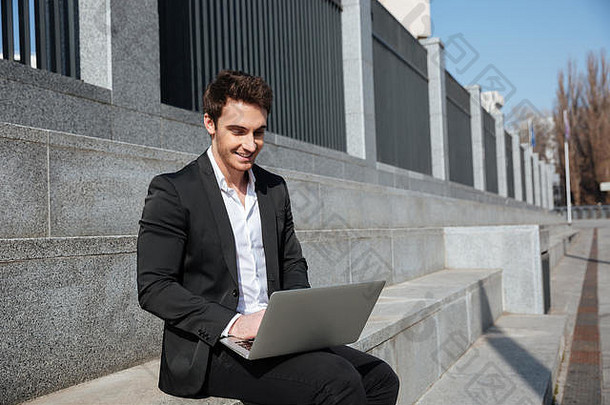 照片微笑年轻的商人坐着在户外移动PC电脑
