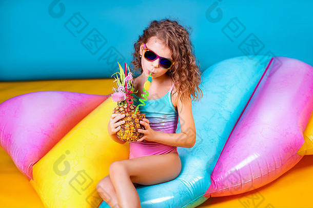 漂亮的微笑女孩穿粉红色的蓝色的泳装太阳镜持有<strong>菠萝</strong>鸡尾酒色彩斑斓的吸管显示拇指彩虹充气床垫背景