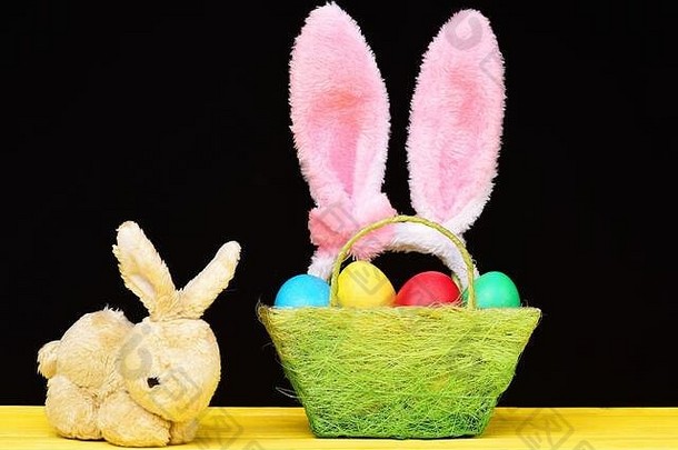 复活节兔子<strong>玩具</strong>绿色篮子站黄色的木表格画鸡蛋红色的蓝色的黄色的绿色颜色粉红色的兔子耳朵希望孤立的黑色的背景假期概念