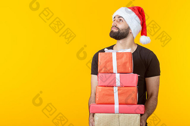 戴着圣诞老人帽子、留着胡子、心怀不满的年轻人拿着五个礼盒，在黄色背景上摆着姿势，并留有文字空间。礼物和礼物的概念