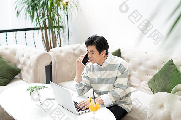 一位亚洲商人在咖啡馆用笔记本电脑工作时使用手机