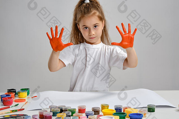 穿着白色t恤的小女孩坐在桌旁，手里拿着惠特曼和五颜六色的颜料，展示着她画过的手。白色的隔离带。中等特写镜头。