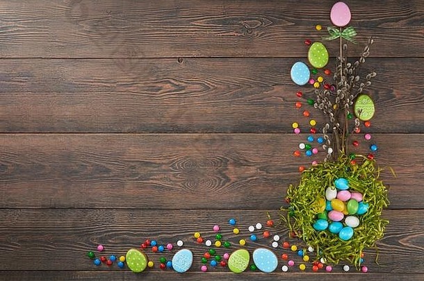 从上面看，小花<strong>生巧</strong>克力球在糖壳里，柳枝孤立在木质背景上。绿色鸟巢里五颜六色的复活节彩蛋，附近有饼干。春季和复活节假期的概念。
