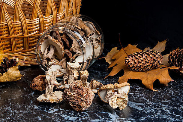 在玻璃罐和桌子上放上干的白色野生蘑菇。背景为黄色木篮，几片干燥的橡树叶、橡子和冷杉球果。