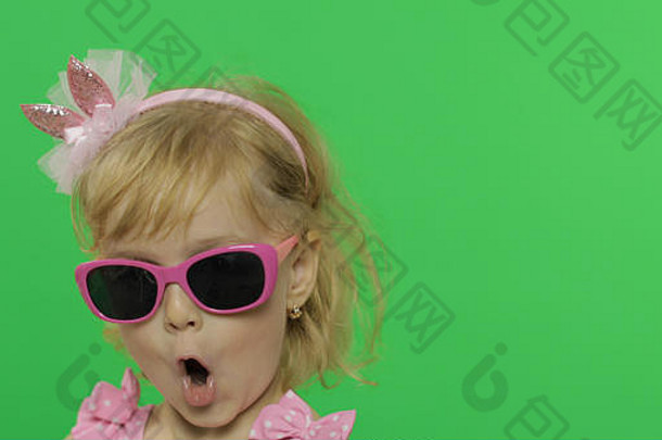 一个积极的女孩穿着粉红色的泳衣，戴着墨镜，对着镜头激动地唱歌。肖像特写。快乐可爱的金发小女孩，3-4岁。做鬼脸和微笑。绿色屏幕。色度键
