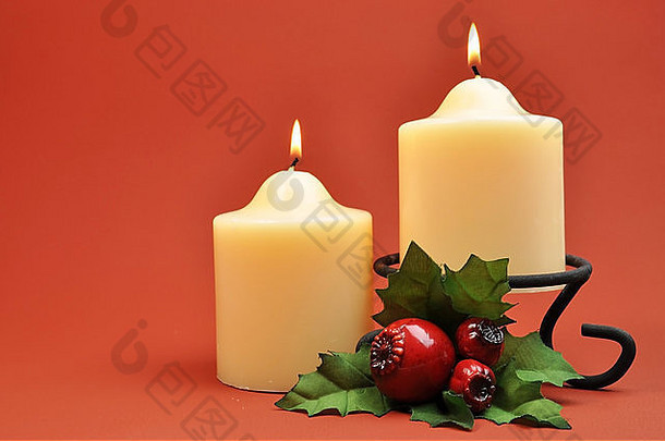 一对乳白色的蜡烛，在红橙色的背景下有绿色和红色的冬青装饰