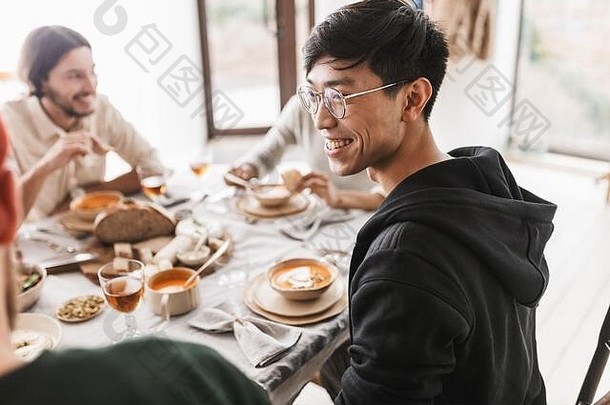 年轻快乐的亚洲男子，深色头发，戴着眼镜，头戴连帽衫，坐在桌旁愉快地向旁边看。<strong>国际友人</strong>小组正在吃午餐